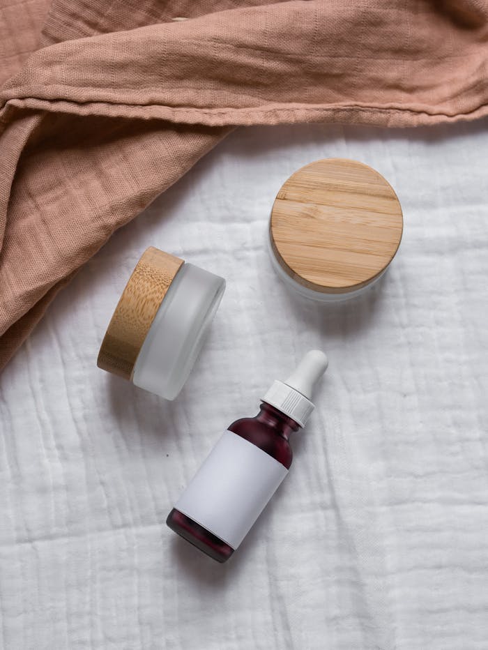 Flatlay of natural skincare jars and dropper bottle with wooden lids on a soft fabric background.